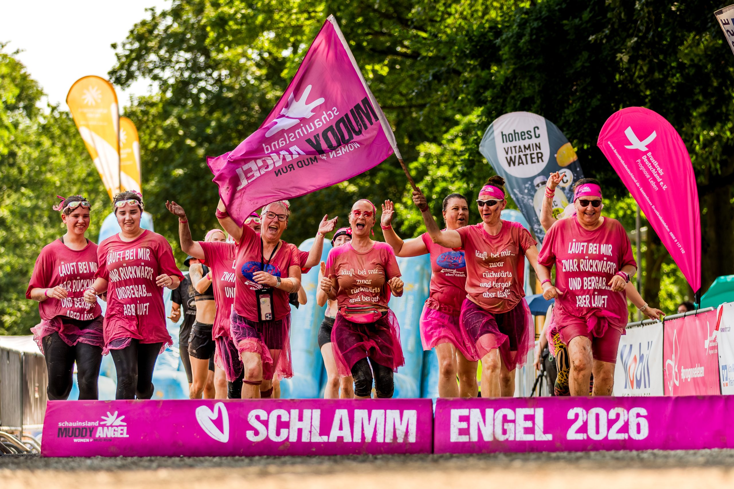 Eine Gruppe schlammverschmierter Teilnehmerinnen läuft jubelnd mit einer Muddy-Angel-Fahne über die Ziellinie.